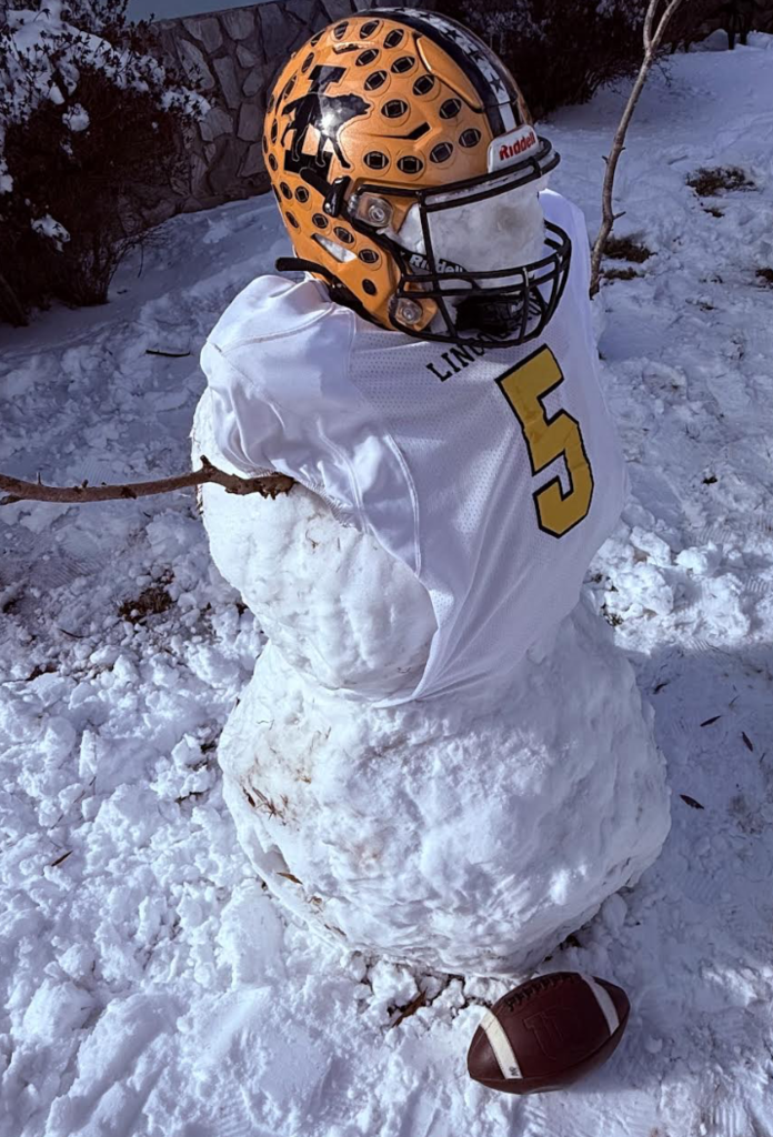 A snowman styled as a football player wearing a Lincoln football jersey and helmet stands in the snow with a football at its base and stick arms extended.
