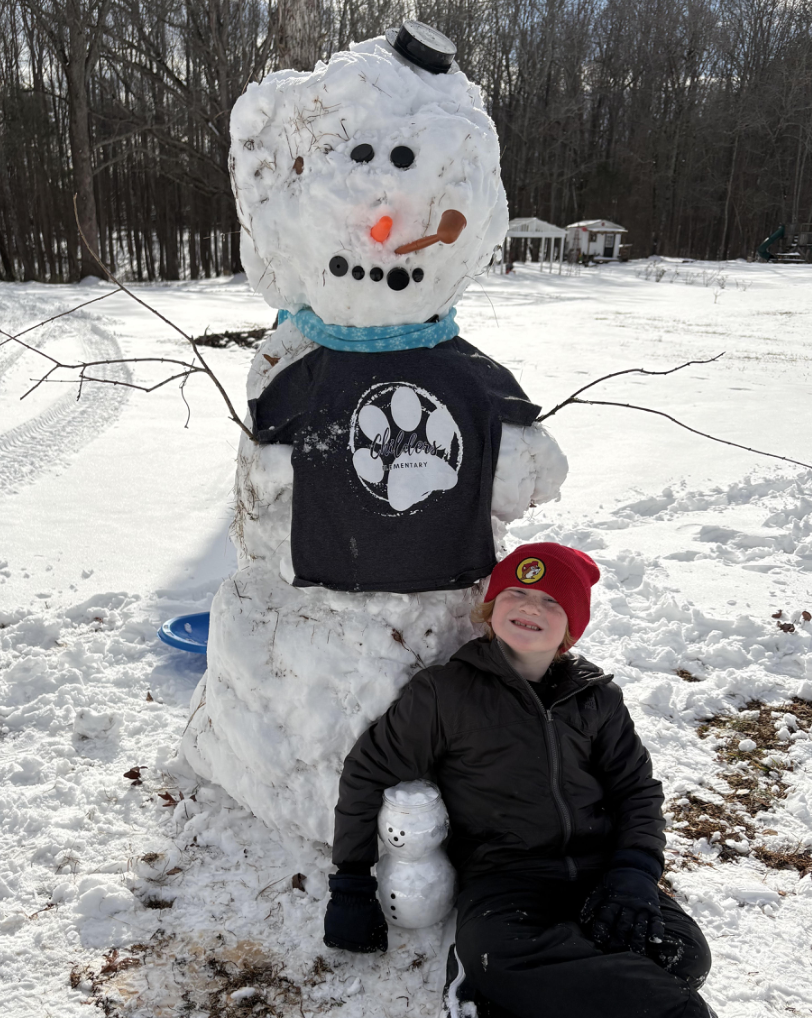 A smiling child in a red winter hat sits beside a tall snowman wearing a school T-shirt, holding a tiny matching snowman in their lap on a bright winter day.