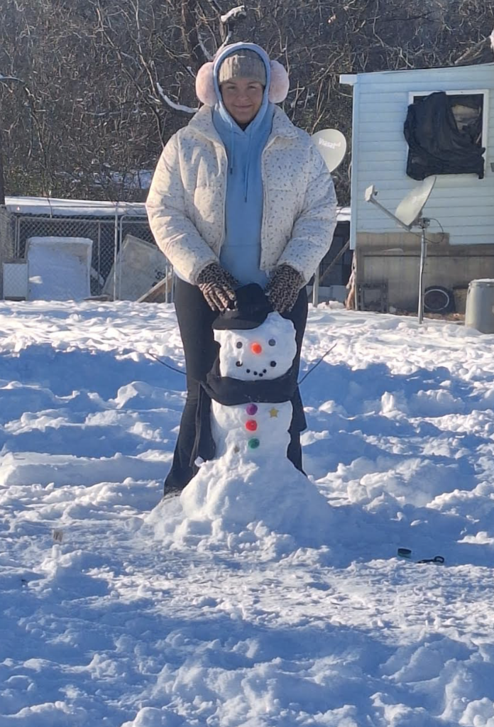 A student wearing earmuffs and a winter coat hugs a small, cheerful snowman decorated with colorful buttons, a scarf, and a black hat in a sunny, snow-covered yard.