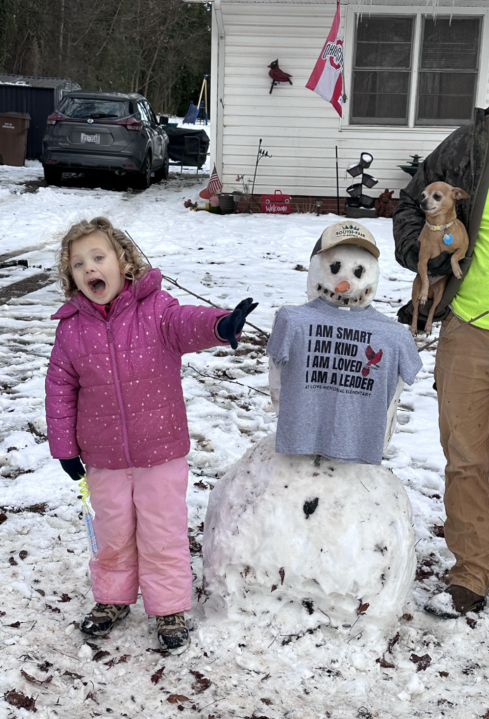 A young student in a pink winter coat stands next to a snowman wearing a gray “I Am Smart, I Am Kind, I Am Loved, I Am a Leader – Love Memorial Elementary” T-shirt. The child reaches toward the camera with an excited expression.