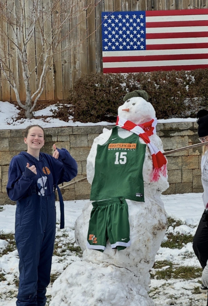 A student gives two thumbs up beside a snowman dressed in an East Lincoln basketball jersey and shorts, complete with a festive scarf in a snowy backyard.
