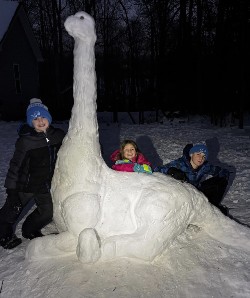 Three children smile beside a giant snow sculpture shaped like a dinosaur with a long neck. The kids are bundled in winter coats and hats, posing proudly with their creative snow creation at dusk.