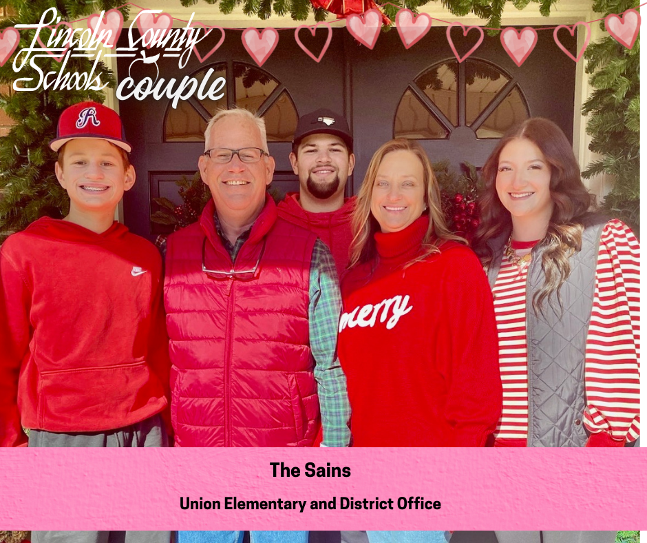 Family photo labeled “The Sains” featuring five people standing together in front of a decorated doorway with holiday greenery. All are dressed in red and festive clothing and smiling at the camera. A string of pink hearts and a Lincoln County Schools Couple graphic appear at the top, and a pink caption bar reads “The Sains – Union Elementary and District Office.”
