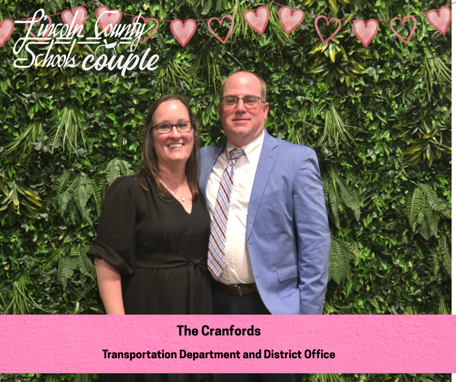 A couple labeled “The Cranfords” stands together smiling in front of a green leafy backdrop. The woman wears a black dress and glasses, and the man wears a light blue suit jacket with a patterned tie. A string of pink hearts and a Lincoln County Schools Couple graphic appear at the top, with a pink caption bar reading “The Cranfords – Transportation Department and District Office.”