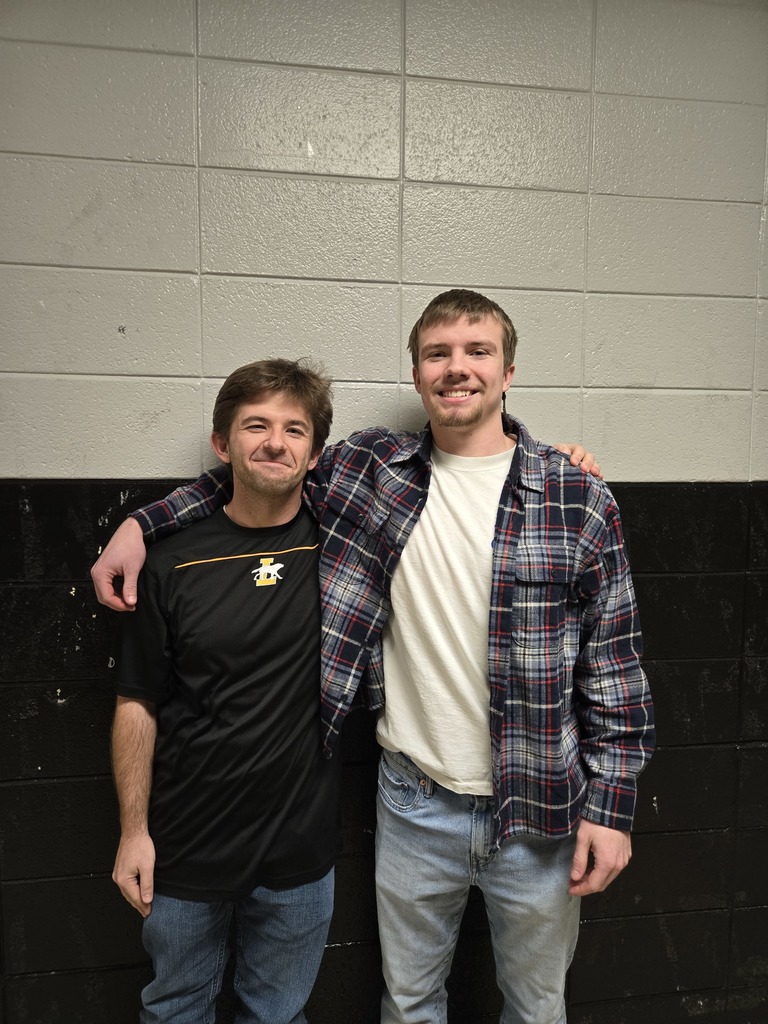 Two young men stand smiling side by side in front of a black and gray block wall. The man on the left wears a black athletic shirt with a small school logo, and the man on the right wears a white shirt with an open plaid flannel. The taller man has his arm draped around the other’s shoulder as they pose casually together.