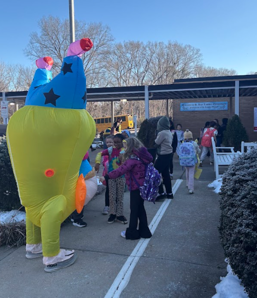 Students gather around the inflatable upside-down clown character outside the school entrance as they arrive in the morning. A school bus is visible in the background and children smile, talk, and reach out to interact with the costume.