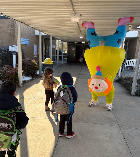 Young students approach the inflatable upside-down clown under a covered walkway at school. Children wearing winter coats and backpacks pause to look at the colorful character while arriving for the day.