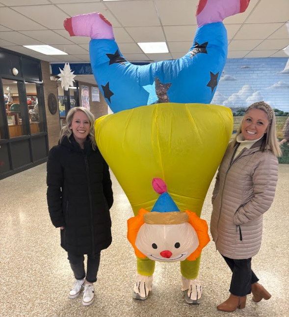 Two smiling staff members stand beside a person wearing an inflatable clown costume in a school hallway. The costume shows the clown upside down doing a handstand, with bright yellow pants, a blue shirt with black stars, pink shoes, and an orange-haired clown face near the floor. A paper snowflake hangs from the ceiling behind them.
