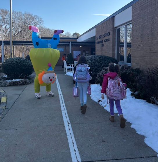 An adult in the inflatable upside-down clown costume greets students outside an elementary school entrance. Two students wearing backpacks walk toward the building along a sidewalk with small patches of snow beside it.