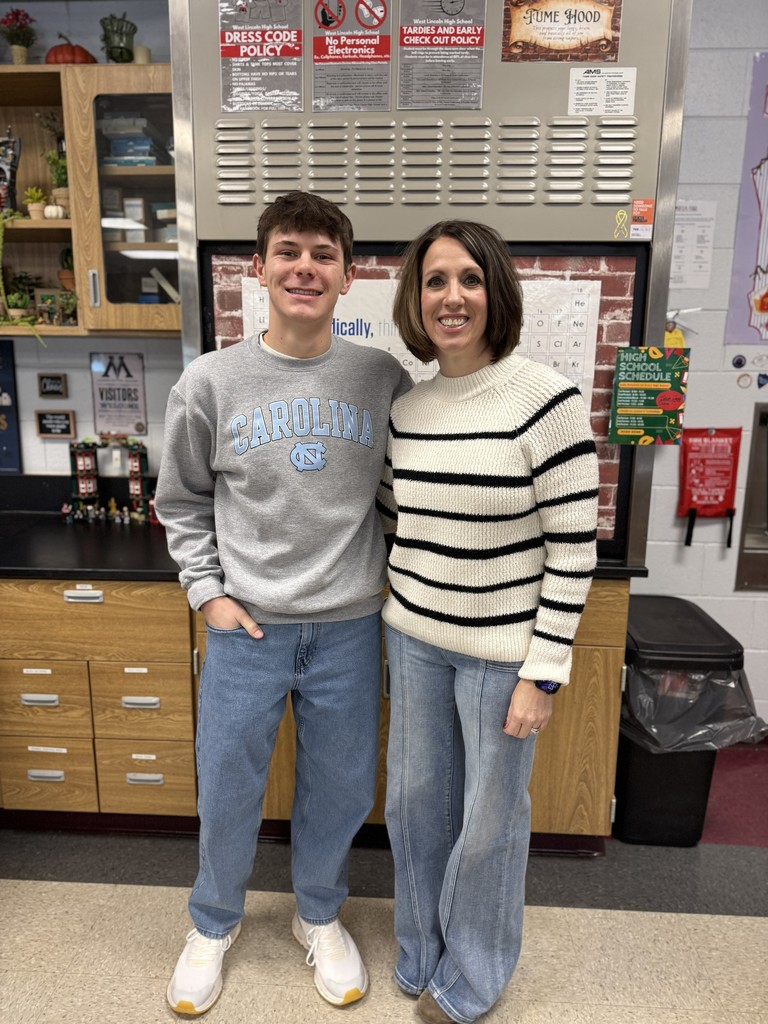 A smiling teenage boy and a woman stand side by side in a classroom. The student wears a gray “Carolina” sweatshirt, jeans, and white sneakers with his hand in his pocket. The woman wears a cream sweater with black horizontal stripes, jeans, and a watch. Behind them are classroom cabinets, a periodic table poster, and several school policy signs posted on metal lockers.