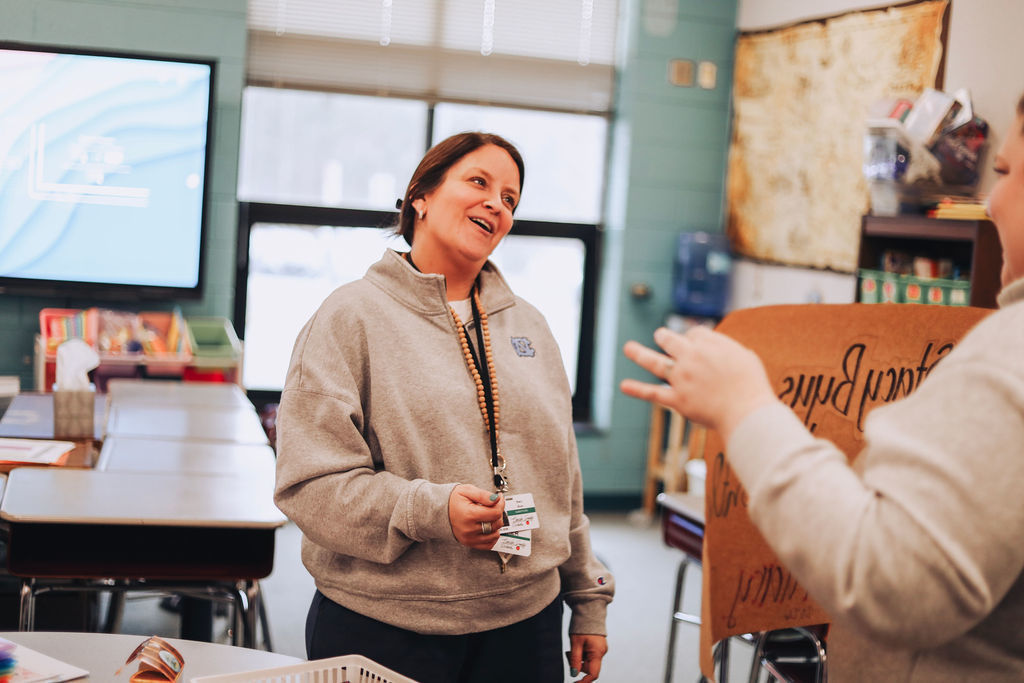 A substitute teacher smiles while speaking with a colleague who holds a recognition poster in a classroom setting.