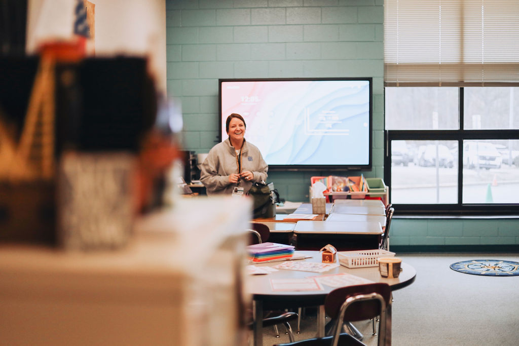 A substitute teacher stands smiling at the front of an elementary classroom with student desks arranged in groups, a large screen on the wall behind her, and daylight coming through the windows.