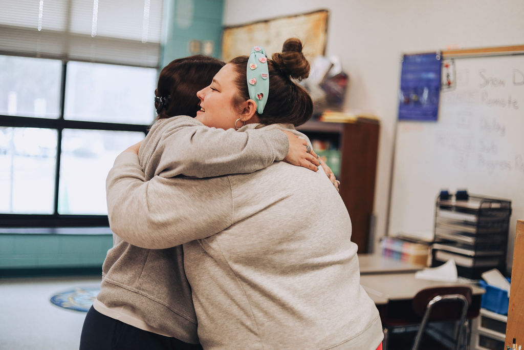 Two women embrace in a classroom, celebrating recognition as colleagues look on and classroom materials are visible in the background.