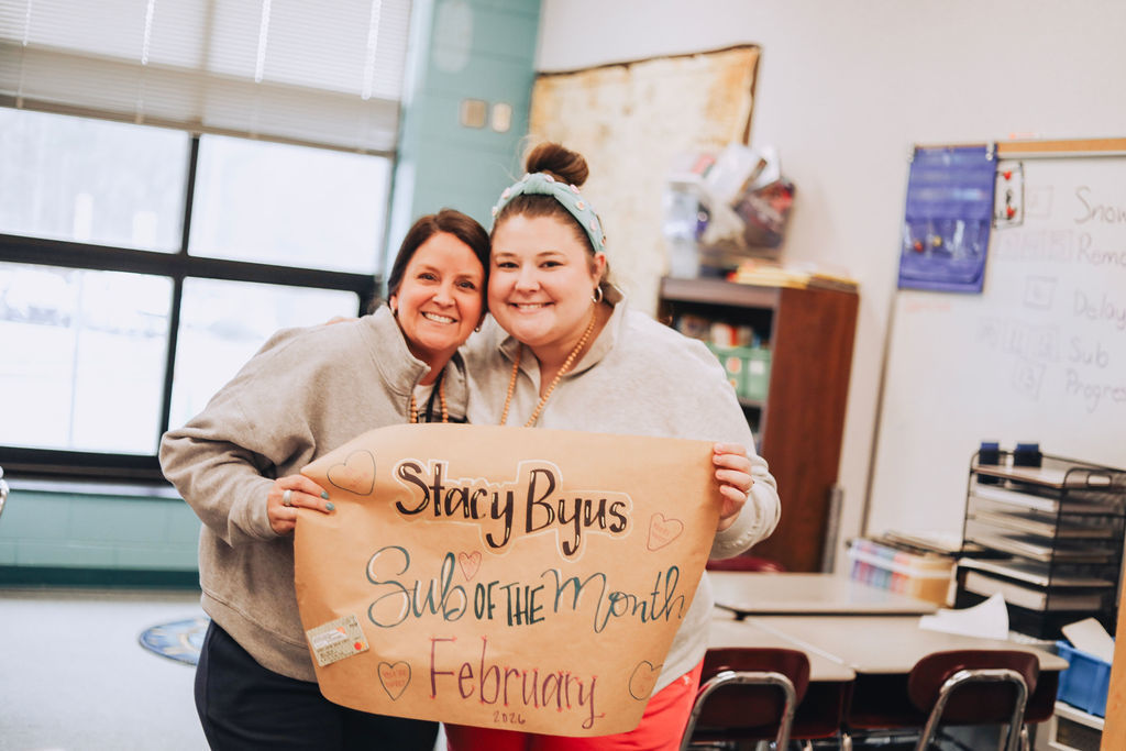 Two women stand side by side in a classroom holding a hand-lettered poster that reads “Stacy Byus Sub of the Month February 2026,” smiling at the camera.