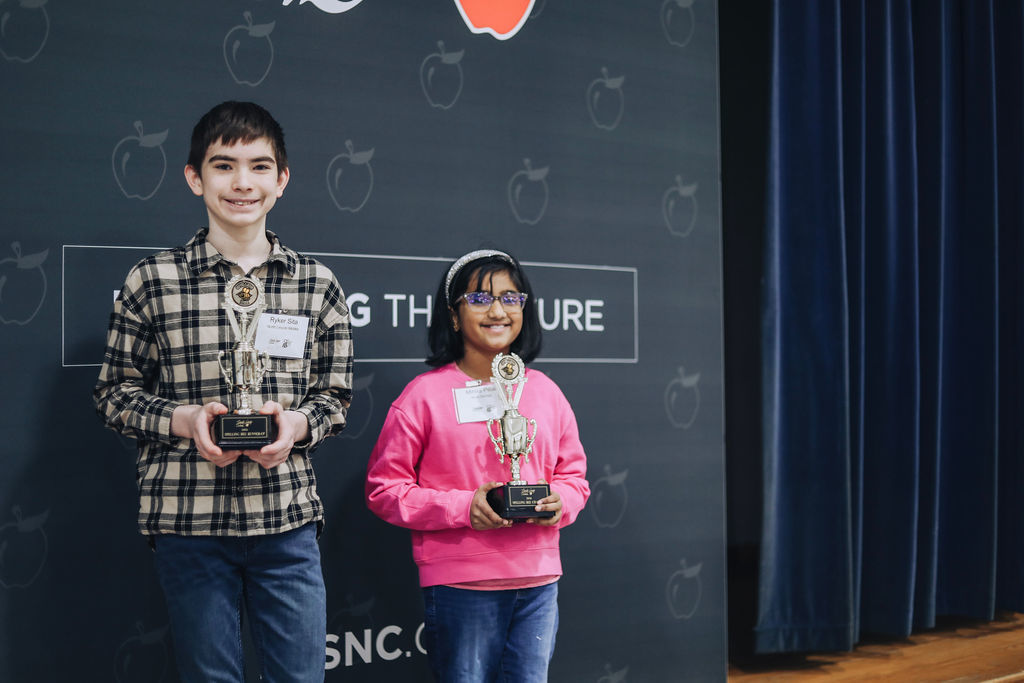 Two students stand on a stage in front of a Lincoln County Schools backdrop holding trophies. The student on the left, Ryker from North Lincoln Middle School, holds a runner-up trophy and wears a plaid shirt and jeans. The student on the right, Mihira from Rock Springs Elementary, holds a first-place trophy and wears a bright pink sweatshirt and glasses. Both are smiling.