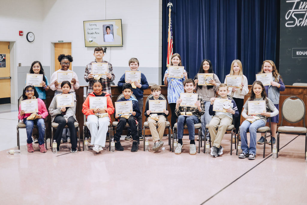 Group photo of elementary and middle school students seated and standing in rows inside a school building, each holding a certificate from the district spelling bee. A screen behind them displays a student’s name and photo, and an American flag stands near the stage.