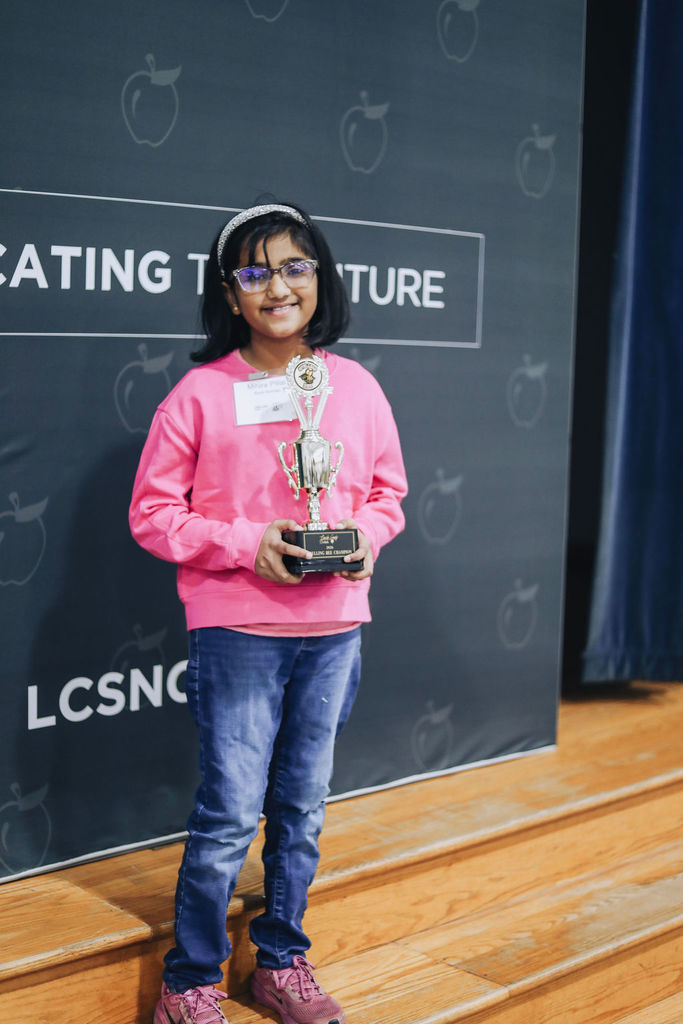 Mihira from Rock Springs Elementary smiles while holding her Spelling Bee Champion trophy. She stands on a wooden stage in front of a Lincoln County Schools backdrop featuring apple icons and district branding.