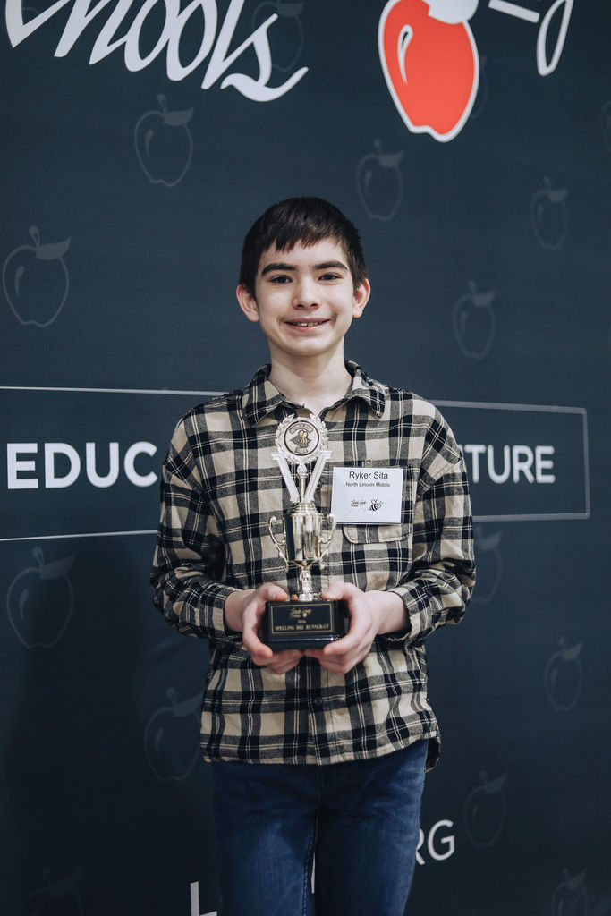 Close-up of Ryker from North Lincoln Middle School smiling and holding his Spelling Bee Runner-Up trophy. He stands in front of a Lincoln County Schools backdrop with apple icons and “Educating the Future” visible behind him.
