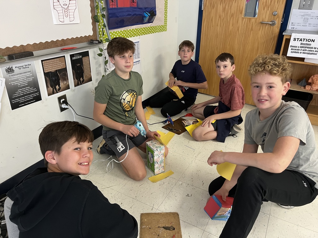 Five North Lincoln Middle School students sit on the classroom floor working together to build and decorate sarcophagus projects from cardboard boxes and colored paper. One student uses a hot glue gun while others hold yellow paper pieces. Classroom posters and supply shelves are visible in the background.
