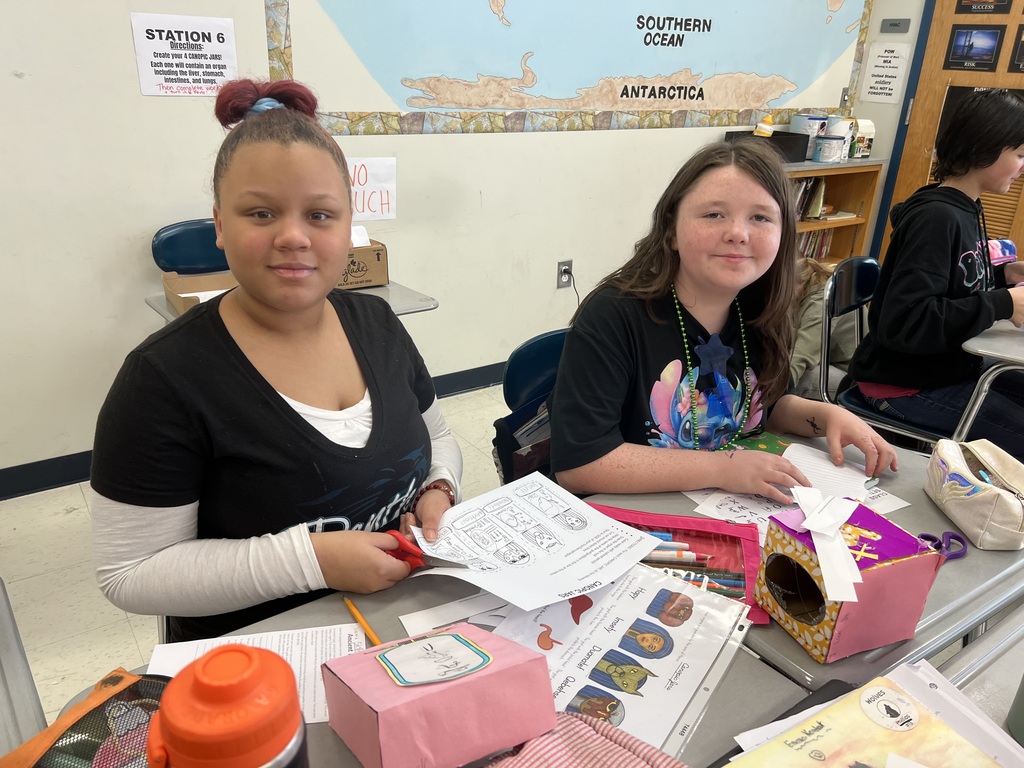 Two students sit at desks cutting paper and assembling colorful sarcophagi projects. One holds a worksheet featuring drawings of Egyptian canopic jars while the other works on a pink decorated box. A classroom world map and storage shelves are visible behind them.