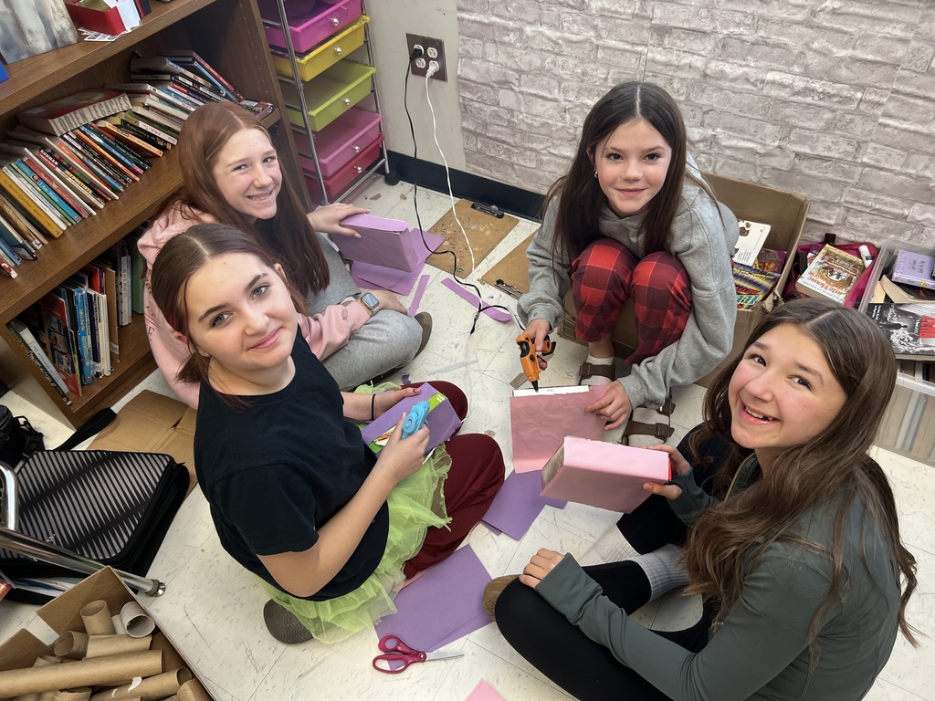 Four students sit in a small group on the classroom floor near a bookshelf, cutting and gluing purple and pink paper onto cardboard boxes to create sarcophagi. Scissors, paper scraps, and craft supplies are spread around them as they smile at the camera.
