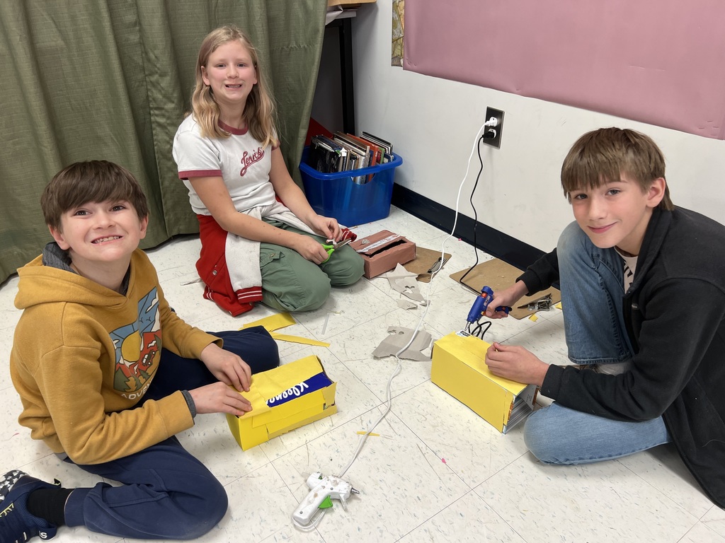 Three students sit on the classroom floor assembling bright yellow cardboard sarcophagi. One student uses a hot glue gun while another presses paper into place. Craft materials and paper scraps are scattered nearby