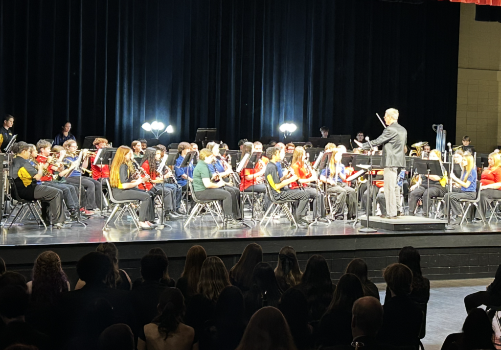 A middle school concert band performs on stage beneath concert lighting. Students wearing red, blue, green, and black school shirts sit in rows with music stands and instruments, including clarinets, flutes, and brass. A conductor stands on a podium leading the ensemble. An audience is seated in the foreground observing the performance.