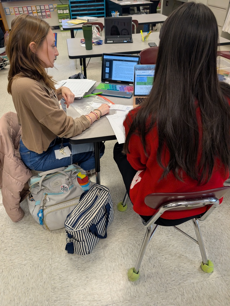 A close-up view from behind a teacher with long dark hair wearing a red sweater, sitting across from a colleague in a tan shirt. They are both looking at laptop screens displaying digital documents and spreadsheets. A large "Be Kind" tote bag sits on the floor next to them.