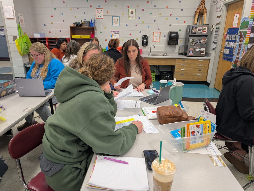 A group of teachers engaged in a collaborative meeting at a long table. They are reviewing paper documents and using laptops. A woman in a rust-colored cardigan looks toward a colleague. The background shows a classroom sink area, a refrigerator with photos, and polka-dot wall decorations.
