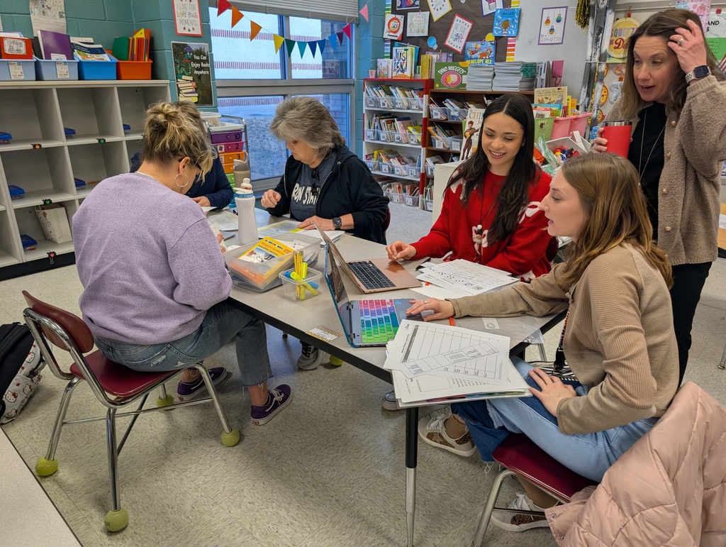 Five female educators sitting and standing around a classroom table during a professional development session. Two women are looking at laptops, while others examine printed documents. The classroom background features bookshelves, a window with colorful bunting, and educational posters.