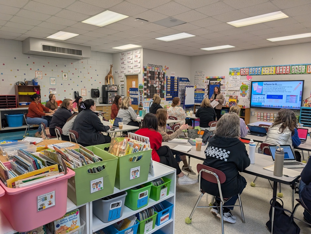 A wide shot of a classroom filled with teachers seated at tables for a presentation. A facilitator stands at the front of the room near a large digital display screen that reads "Where is it..." and shows screenshots of a website. The room is organized with bins of books and educational posters on the walls.