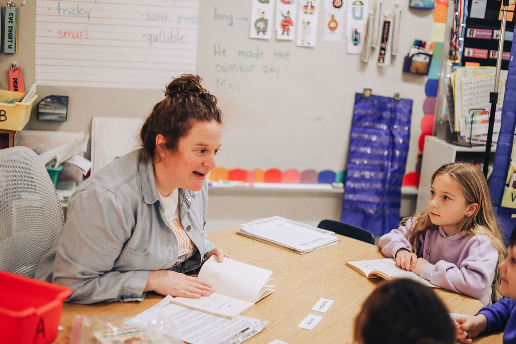 A female teacher with her hair in a bun sits at a wooden table, pointing to a page in a book while speaking to a young girl in a purple hoodie. The background features a whiteboard with handwritten vocabulary words like smart and gullible.