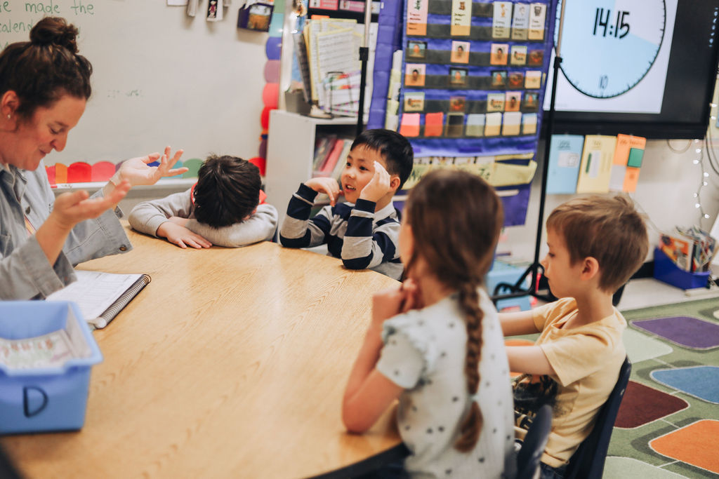 A teacher gestures with both hands while sitting at a table with four students. One student has their head down on the table, another is looking at the teacher with hands near their face, and two others sit with their backs to the camera. A digital clock on a screen in the background shows the time 14:15.