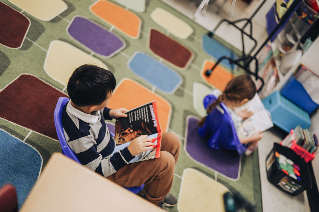 High-angle view of two students sitting in blue plastic floor chairs on a green rug with colorful rectangular patterns. Both students are focused on reading their own books.