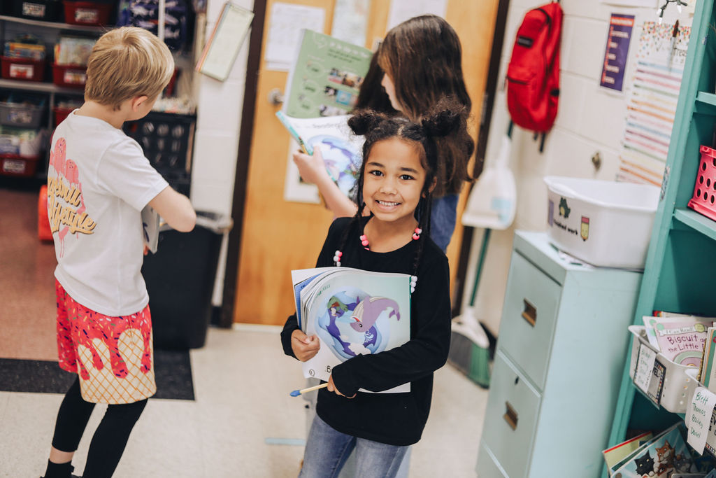 A young girl with her hair in two buns smiles at the camera while holding a stack of books with a whale illustration on the cover. Other students are in the background of the classroom, which includes a teal bookshelf and a red backpack hanging on the wall.