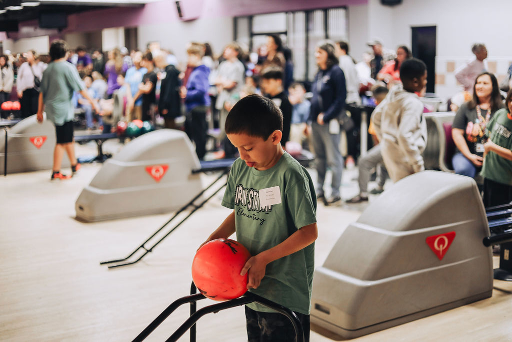 A young boy wearing a green Catawba Springs Elementary t-shirt and a name tag stands at the top of a bowling lane. He is looking down intently as he holds a bright red bowling ball with both hands, preparing to place it on a black metal bowling ramp.