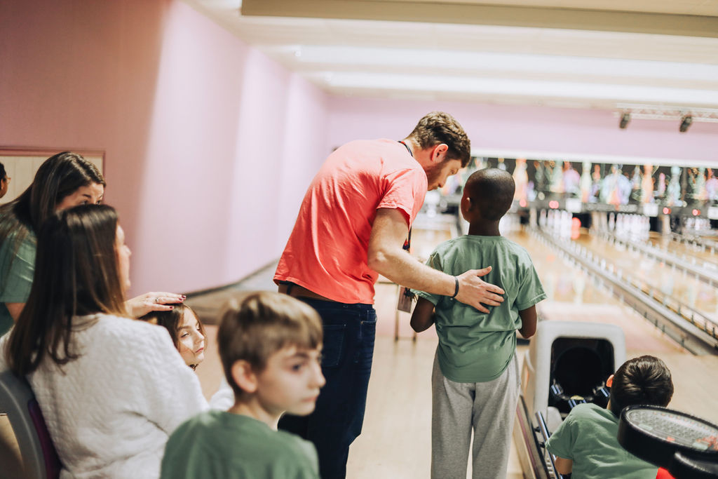 A male volunteer in a red t-shirt leans down to speak to a student in a green shirt at a bowling alley. The volunteer has his hand on the student's back in a supportive gesture. Other students and adults are seated in the foreground, watching the activity on the lanes.