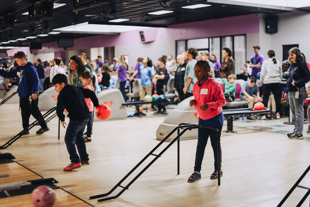 A busy bowling alley scene during a Special Olympics event. In the foreground, a young girl in a bright pink hoodie stands near a black bowling ramp. To her left, a boy in a black hoodie leans forward to watch his red bowling ball roll down the lane. Many other students and adults are visible in the background.