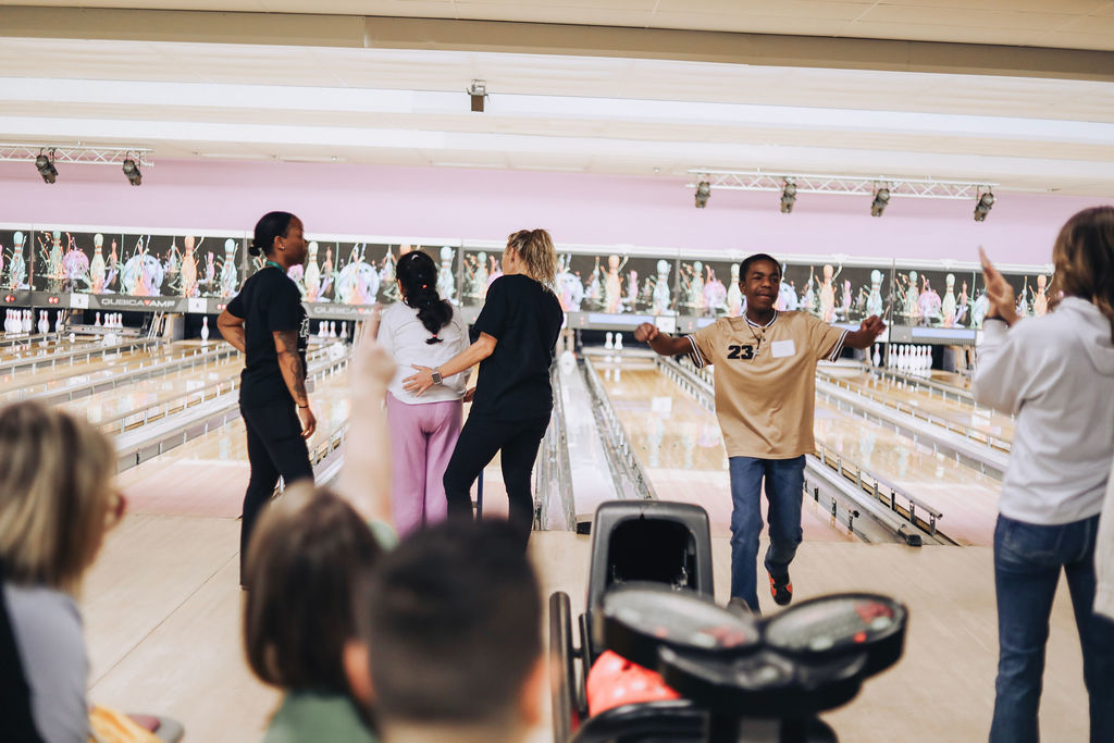 A student in a tan jersey with the number 23 cheers with his arms outstretched while walking back from the bowling lane. In the background, teachers and volunteers stand by the lanes, one with a hand on a student's back, while pins are visible at the end of the multiple lanes.
