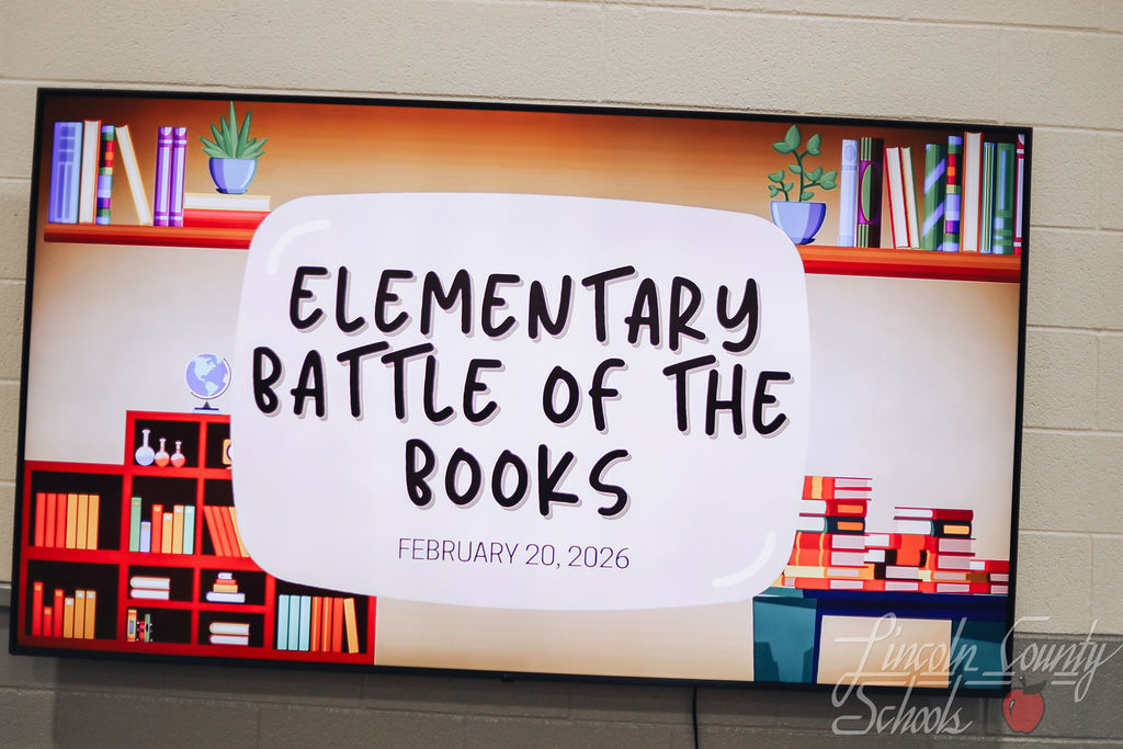 A large digital display screen shows a colorful graphic reading “Elementary Battle of the Books” with the date February 20, 2026. The design features illustrated bookshelves filled with books and small plants on both sides.