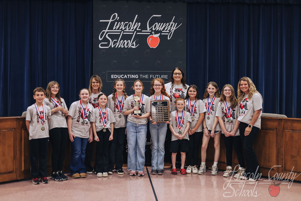 Students from Union Elementary wearing matching gray team shirts stand on stage with medals around their necks, holding a championship plaque. Several adult coaches stand with them. The Lincoln County Schools “Educating the Future” backdrop is displayed behind the group.