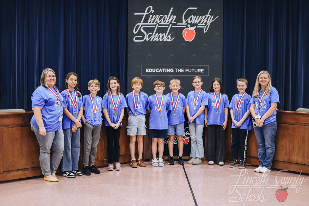 Another elementary school team in matching blue Battle of the Books shirts stands on stage with medals around their necks. Two adult coaches stand at the ends of the group. The Lincoln County Schools “Educating the Future” backdrop is visible behind them.
