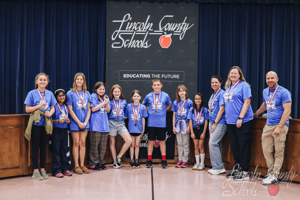 A group of elementary students wearing matching blue Battle of the Books shirts stand on a stage with medals around their necks. They are joined by two adult coaches. Behind them is a Lincoln County Schools backdrop with the phrase “Educating the Future.”