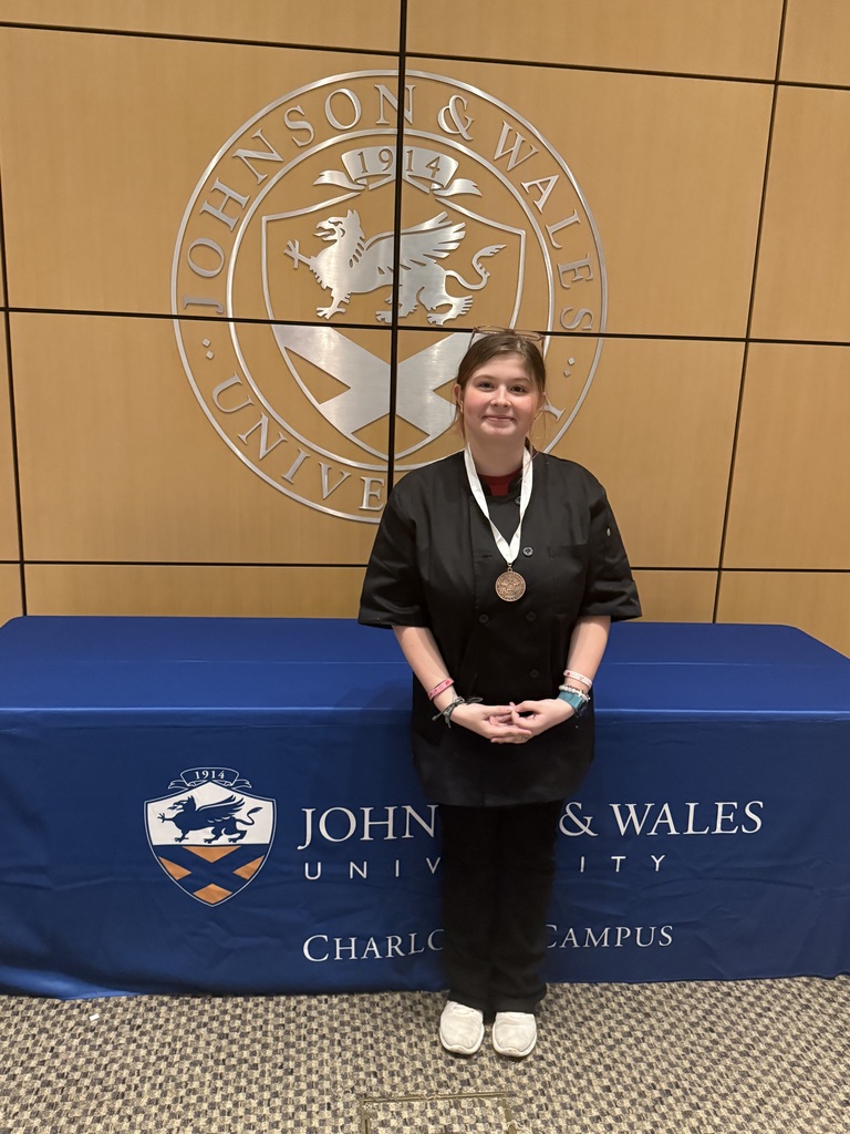 A student wearing a black chef’s jacket stands in front of a Johnson & Wales University backdrop with a university seal displayed behind her. She is wearing a bronze medal around her neck and stands with hands clasped in front of her, smiling proudly. A blue tablecloth in front of her displays the Johnson & Wales University logo and “Charlotte Campus.”
