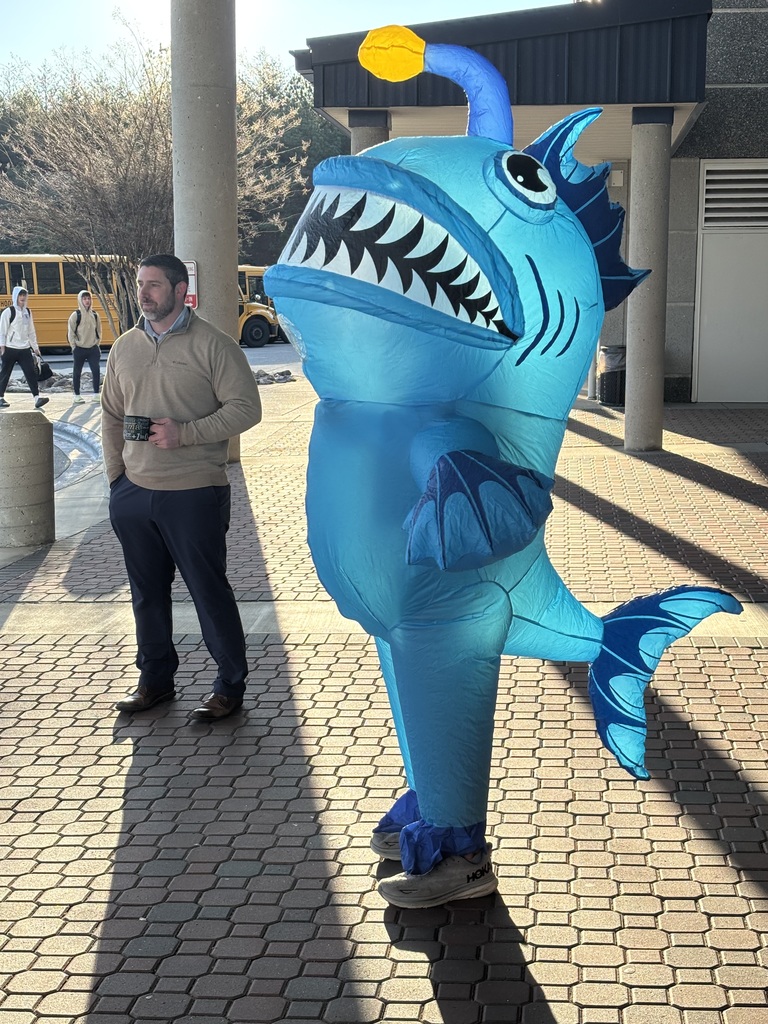 A staff member wearing an inflatable blue anglerfish costume stands outside a school entrance near the bus loop. The costume features large white teeth and a yellow light on top. The staff member stands beside another adult holding a coffee mug while school buses and students arriving for the day are visible in the background.