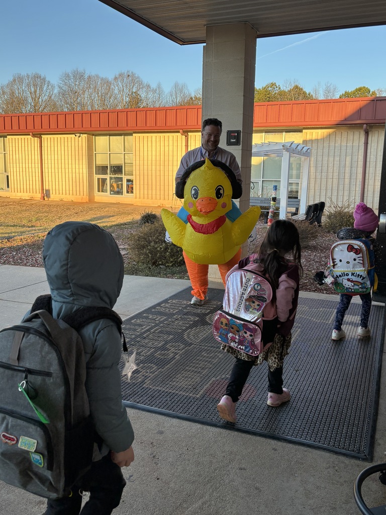 A staff member wearing an inflatable yellow duck costume with black earmuffs stands under a school awning greeting three elementary students as they arrive in the morning. The children, wearing winter coats and colorful backpacks, walk toward the entrance on a covered walkway. The school building with red trim and classroom windows is visible in the background in early morning light.