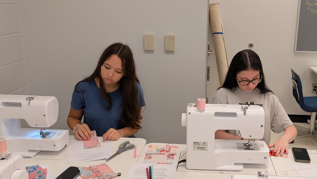 Two students sit at tables using white sewing machines in a classroom. They carefully sew small pink and patterned fabric squares while scissors, thread, and printed instructions are spread across the table.