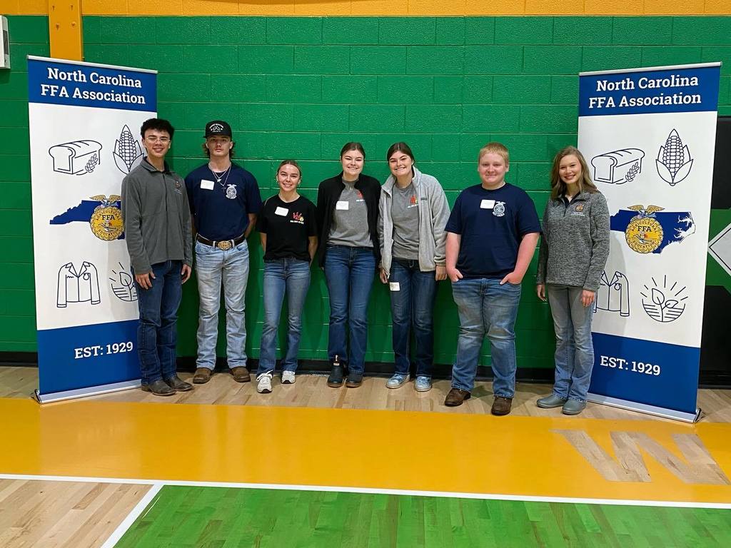 Seven students stand between two North Carolina FFA Association banners inside a gymnasium. The students are dressed in jeans, boots, and FFA attire and smile for a group photo.
