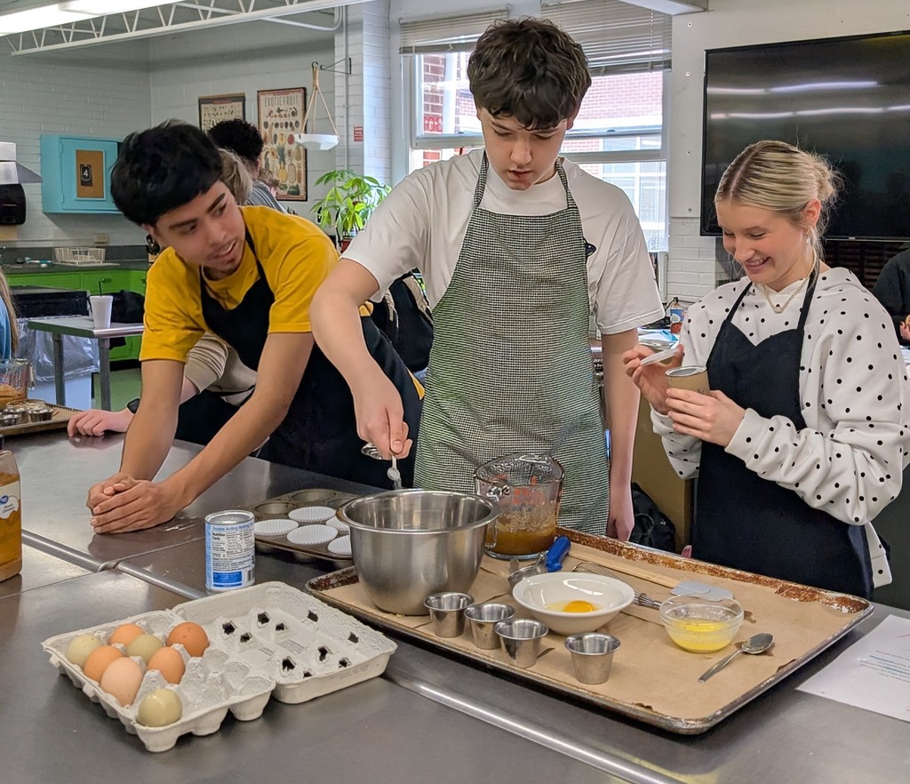 Three students wearing aprons work together in a culinary classroom. One student measures ingredients into a metal mixing bowl while another smiles and holds a small container. Eggs, measuring cups, and baking supplies are arranged on a tray.