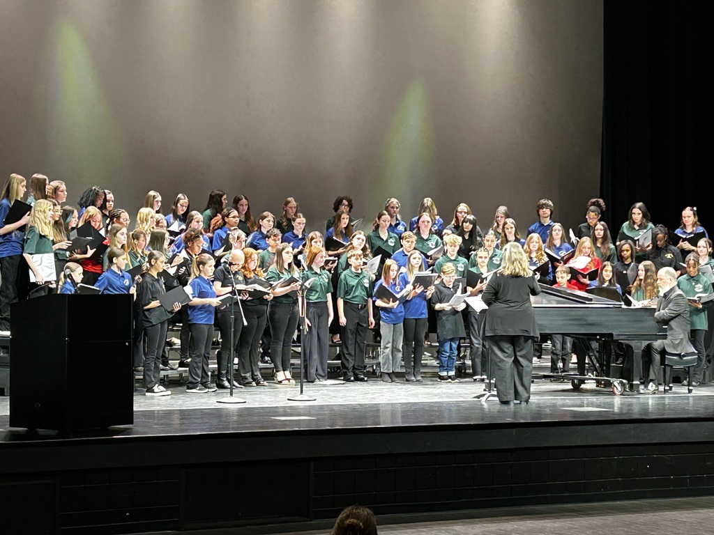 A large middle school honors chorus stands on tiered risers on stage at the Citizens Center. Students wear coordinated black, green, and blue concert attire and hold black music folders while singing. A conductor stands at the front facing the group, and a pianist accompanies them on a grand piano. Stage lighting casts soft green tones on the backdrop.